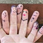 Foto einer Hand vor Backsteinwand, auf deren Fingerspitzen mit Marker verschiedene Gesichter und Accessoires gezeichnet sind.