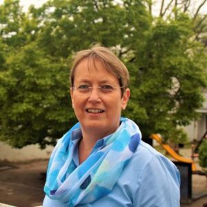 Foto von Roswitha Frenzel mit kurzem, hellbraunem Haar, Brille und blauem Schal vor grünem Baum und gelber Rutsche draußen.