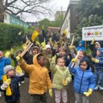 Foto von mehreren Kindern in bunten Jacken und gelben Handschuhen vor der KiTa Rokoko mit Stöcken und gelben Fahnen.