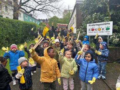Foto von mehreren Kindern in bunten Jacken und gelben Handschuhen vor der KiTa Rokoko mit Stöcken und gelben Fahnen.