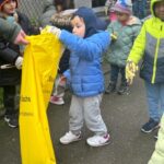 Foto von mehreren Kindern in bunten Winterjacken und Handschuhen, die Müll in einen großen gelben Sack einer erwachsenen Person legen.