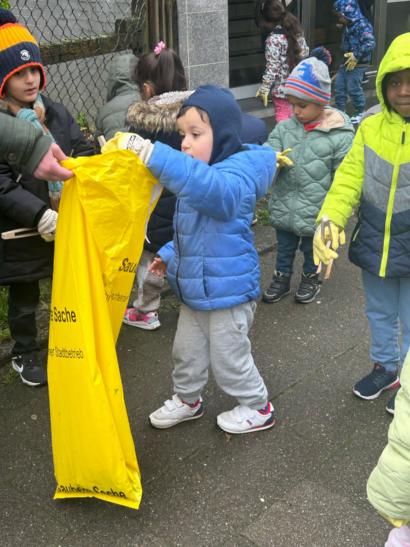 Foto von mehreren Kindern in bunten Winterjacken und Handschuhen, die Müll in einen großen gelben Sack einer erwachsenen Person legen.