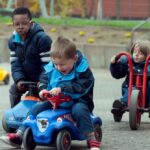 Foto von drei Jungen, die draußen in Jacken fahren: Zwei auf blauen Rutschautos, einer auf einem roten Dreirad.