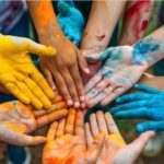 Foto von mehreren Händen, bemalt mit gelber, oranger und blauer Farbe, die in der Bildmitte mit offenen Handflächen zusammenliegen.
