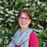 Foto von einer Frau mit kurzen braunen Haaren, roten Brillen, rotem Oberteil und blauem Schal vor grünem Busch mit weißen Blüten.