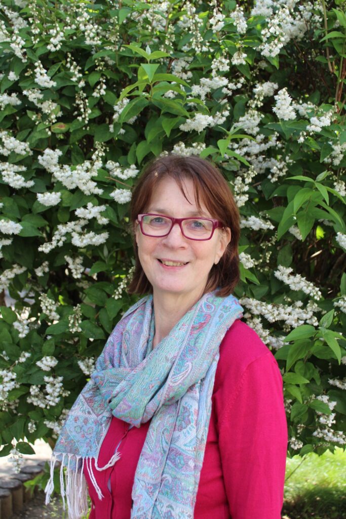 Foto von einer Frau mit kurzen braunen Haaren, roten Brillen, rotem Oberteil und blauem Schal vor grünem Busch mit weißen Blüten.
