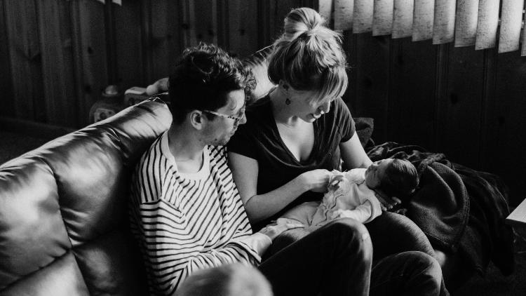 Foto von einem Mann und einer Frau auf einem Sofa. Die Frau hält ein Neugeborenes im Arm. Natürliches Licht fällt in den Raum.