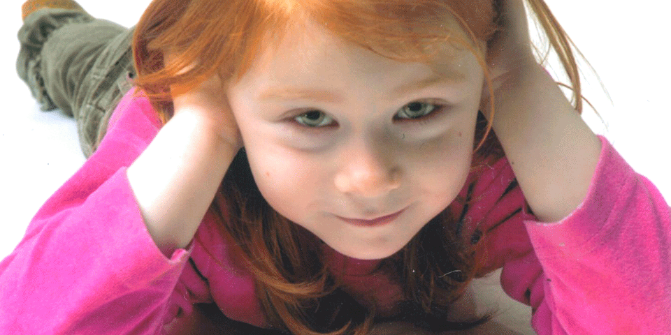 Foto von einem Kind mit roten Haaren und rosa Shirt, das auf dem Boden liegt und in die Kamera lächelt, Kopf in den Händen.