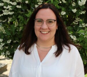 Foto von Roswitha Frenzel mit langen braunen Haaren und Brille vor grünem Busch mit weißen Blüten, trägt weiße Bluse und Silberkette.