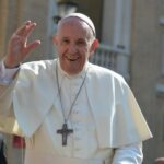 Foto von einem Mann in weißen religiösen Gewändern und weißer Kappe, mit Kreuzkette, winkend vor einem säulengeschmückten Gebäude.