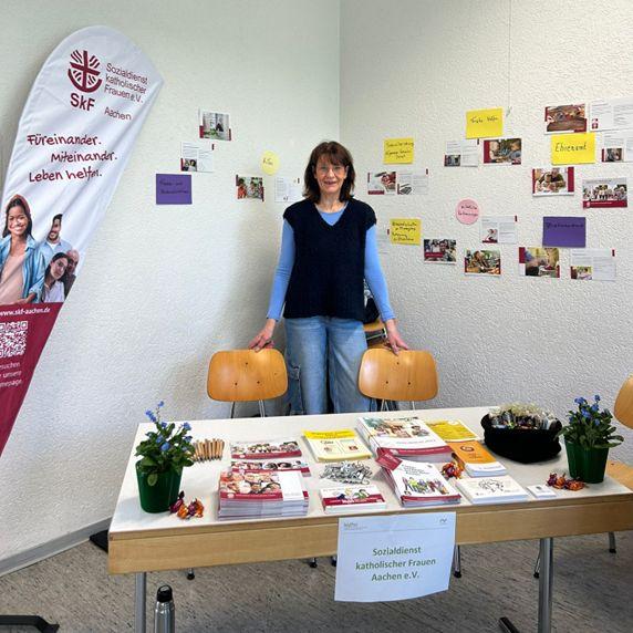 Foto von einer Frau hinter einem Tisch mit Infomaterial und Pflanzen, Banner im Hintergrund, Schild: SkF - Sozialdienst katholischer Frauen Aachen e.V.