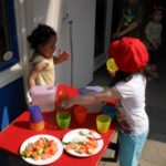 Foto von zwei Kindern, die an einem roten Tisch im Freien spielen. Bunte Becher und Gemüsestücke auf Tellern sind zu sehen.