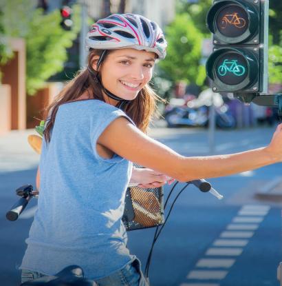 Foto von einer jungen Frau mit Helm, die an einem Fahrradverkehrslicht in Aachen bei Sonne am Zebrastreifen ihr Fahrrad hält.